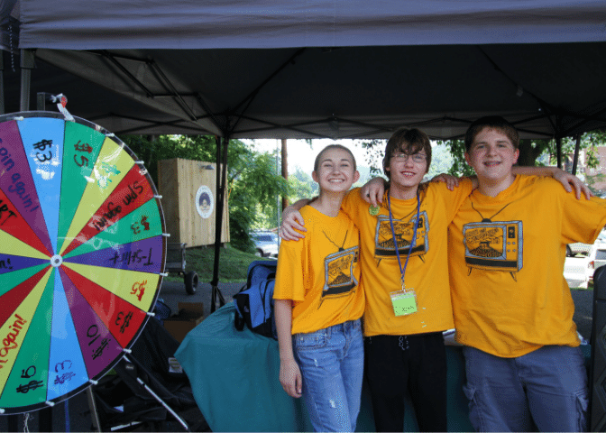Three people stand next to a wheel at letcher county farmers market. The market supports local farmers and artists in eastern kentucky